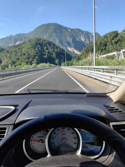 The image shows a view from inside a car driving on a highway. The dashboard and steering wheel are in the foreground, with the road stretching out ahead towards a mountainous landscape. The sky is clear and blue. There are guardrails on either side of the highway, and streetlights are visible in the distance. The mountains are covered in lush green trees. A white structure, possibly a bridge or an elevated walkway, is visible on the right side of the frame. The car's speedometer shows a speed of approximately 60 km/h. The scene appears to be set in a rural or semi-rural area, given the natural scenery. The location is identified as Adlerskiy, Russia.