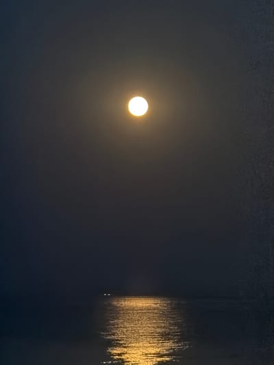 The image depicts a nighttime scene over a body of water, likely the sea, near Νέα Καλλικράτεια, Greece. A bright, full moon hangs in the dark sky, casting a shimmering golden reflection onto the water's surface. The reflection extends from the horizon upwards, creating a luminous path. In the distance, two small, indistinct lights are visible on the water, possibly from a boat or a distant structure. The overall atmosphere is serene and tranquil.
