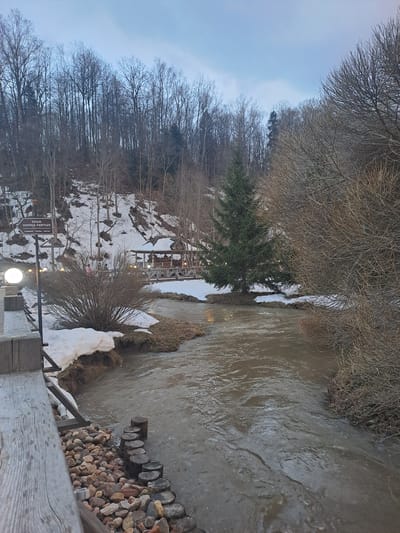 The image depicts a landscape in what appears to be a partially snow-covered area. A body of water, possibly a river or stream with murky water, flows through the scene. The riverbank is lined with vegetation, some of which is bare due to the season. Patches of snow are visible along the banks and on the surrounding landscape. Bare trees and branches dominate the left side of the frame. In the distance, what appears to be a building or structure, possibly a restaurant, is visible on a slope. A wooden railing and rocks are present in the lower right corner, suggesting a viewpoint or pathway. The sign present reads, "Храм Добрка Овитх". The location is Vzglyadnevo, Russia, based on the sign's context.