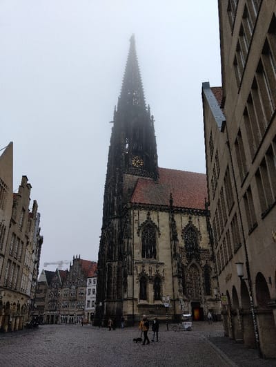 This image depicts a street scene on a foggy, overcast day in Münster, Germany, specifically featuring the St. Lamberti Church at the Prinzipalmarkt. The prominent structure is the Gothic St. Lamberti Church, centrally positioned, displaying a dark, ornate stone facade, a red-tiled roof, and a tall, slender spire. The spire's uppermost section is obscured by dense fog, while a golden clock face on the tower indicates approximately 11:20.
To the left of the church, a row of historic, multi-story buildings with stepped gables and arched ground-floor facades lines the cobblestone street. To the immediate right, a light beige, multi-story building with vertical window patterns features an arched arcade at its base, where a streetlamp is affixed. The cobblestone square in the foreground and mid-ground appears damp.
Approximately 6-8 pedestrians are visible. In the mid-ground, a person in a yellow jacket walks a small, dark dog on a leash, accompanied by another individual in a dark coat. A white bicycle and a small white vendor cart, marked "Doni," are parked near the church's entrance. In the background, further down the street, additional gabled buildings are visible, with a construction crane extending above them against the uniformly gray, foggy sky. Various street signs are present near the church.
