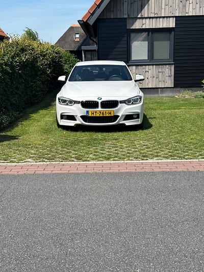 In Ossenzijl, Netherlands, a white BMW sedan is parked on a grassy driveway bordered by a red brick walkway and asphalt road. The car faces the viewer, with its license plate reading "HT-761-H". Behind the car, a dark-colored modern house with a wooden facade and a thatched roof is visible. A neatly trimmed green hedge lines the left side of the driveway. The sky is clear and blue.