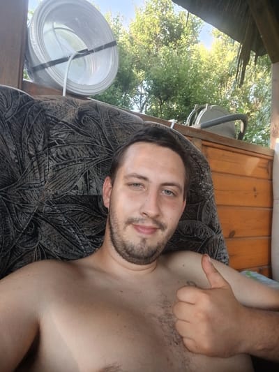 The image features a young man lying down, possibly on an outdoor sofa or bench. He is shirtless, revealing his chest and shoulder. The man has short brown hair, a beard, and blue eyes. His facial expression is neutral. His right hand is raised, making a "shaka" gesture.
The setting appears to be outdoors, with visible elements of nature including green trees and a bright blue sky. There's a wooden structure in the background, suggesting a porch, balcony, or shed. A roll of white material, possibly cable or tubing, is visible to the right of the man, wrapped in plastic and secured with black tape.
The lighting suggests it is daytime, likely a sunny day, as indicated by the bright sky and the dappled light filtering through the trees. There is no visible text in the image. The overall impression is one of a relaxed, casual moment in an outdoor, possibly rural, environment.