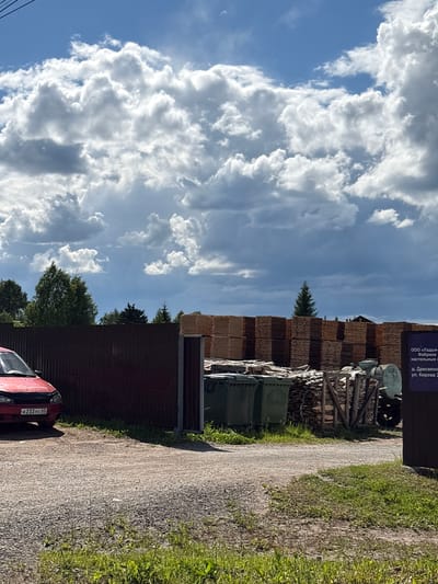 The image depicts a rustic outdoor scene in Дресвяново, Russia, likely during the daytime with a blue sky filled with dramatic, billowing white clouds. In the foreground, a red car with a license plate reading "A 233 YC 43" is partially visible, parked on a gravel road that curves to the right. To the left of the road, a dark brown metal fence runs along the property.

Beyond the fence, a large pile of stacked lumber is visible, indicating a lumber yard or woodworking facility. Some green industrial bins are also present near the lumber stacks. A wooden structure, possibly part of the lumber yard's infrastructure, stands near the stacks. In the background, lush green trees, including a tall evergreen, punctuate the landscape.

There is visible text on a sign attached to one of the structures, which appears to be related to a company named "ООО «Ладья- Фабрика настольных»" located at "д. Дресвяно, ул. Кирова 2". The overall impression is one of a working industrial area set against a backdrop of natural beauty, with the dynamic cloud formation suggesting potentially changing weather or simply a striking sky. The scene appears calm and business-like, with no people immediately visible.