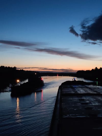 The image depicts a river scene at dusk. In the foreground, a dark, flat surface appears to be part of a barge or dock. To the left, another vessel, possibly a tugboat or barge, is visible with its navigation lights illuminated, creating reflections on the water. The river reflects the fading light of the sunset. The banks of the river are lined with trees. The sky exhibits a gradient of colors, transitioning from deep blue at the top to orange and pink near the horizon. A few clouds are scattered across the sky. There is no evidence in the image to confirm that the location is in the city of Uzhla, Russia.