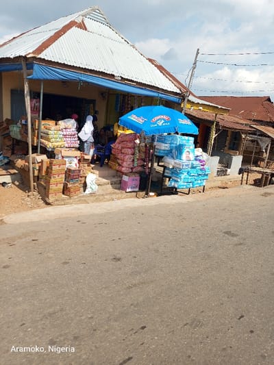 The image shows a small shop with a blue awning in front of it. There are boxes of various goods stacked on the ground and a blue umbrella with a white advertisement is being used as an awning for another stall. There is a small metal rack with a variety of goods on it. The image appears to be taken in front of a shop, in the city of Ilobu, Nigeria, at the coordinates provided. The pavement in front of the shop is a dusty grey and there are a couple of people standing in front of the shop. The shop is located on a road, with other buildings visible in the background. There are power lines running along the top of the buildings, and a small street with a light grey pavement is visible in the background. The sky is a light blue with some wispy white clouds.