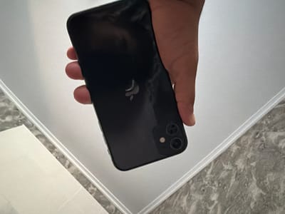 In Avdarma, Moldova, a person's hand is holding a black iPhone, specifically model 11 or 12, against a light-colored surface, possibly a table. The phone's rear camera module, featuring two lenses and a flash, is visible. The iPhone's Apple logo is also discernible on its back. The surrounding environment appears to be a domestic setting, with hints of a countertop or desk. The lighting is bright and diffused, creating soft shadows. The composition is a close-up shot, focusing on the phone and the hand holding it.