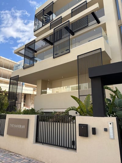 A modern, multi-story residential building named "THE SAMIRAH LUXURY RESIDENCES IN ELLINIKO" is depicted under a clear blue sky with scattered white clouds in the Municipal Unit of Elliniko, Greece. The building features a light beige textured facade, glass-paneled balconies, and prominent black vertical-slat screens, some fixed and others appearing operable. The ground level is secured by a light beige wall and a black vertical-slat metal fence. Mounted on the exterior wall are a rectangular dark taupe sign displaying the building's name, a black wall-mounted mailbox with a decorative wave pattern, a small black rectangular device, a black keypad, and a silver multi-button vertical intercom system. Lush green foliage, including broad-leafed plants and bamboo-like shrubs, is visible around the base of the building and behind the fence. Paved stones are visible in the lower left foreground. No individuals or specific actions are present in the scene.