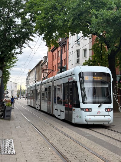 A white modern tram with the text "BOGESTRA" on its side is stopped at a tram stop in Witten, Germany. The tram is on a cobblestone street with tram tracks running through it. Buildings with brick and white facades line the street, and large trees with green leaves provide shade. A person in a blue shirt and dark pants is walking on the sidewalk next to the tram. The sky is overcast.