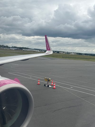 An airport scene is captured from the window of a Wizz Air aircraft, showcasing a wing and engine. The aircraft's left wing extends horizontally across the frame, featuring a distinctive purple wingtip with "wizzair.com" clearly visible. In the bottom right, part of the aircraft's engine, also in purple and white livery, is seen with its fan blades.

The plane is on a grey asphalt tarmac, marked with white lines indicating taxiways or parking positions. Nearby, a yellow piece of ground support equipment and two orange and white traffic cones are visible. Beyond the tarmac, a grassy area and a fence separate it from a distant line of low-rise buildings, likely part of the airport infrastructure or surrounding area. The sky is heavily overcast with dark, dense clouds, suggesting gloomy weather conditions during the daytime. The aircraft appears to be stationary or slowly taxiing on the ground, indicating an activity related to departure or arrival. The location is Tillé, France.