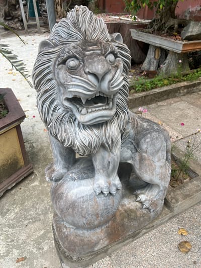 The image depicts an outdoor scene in Đăk Mar, Vietnam, featuring a weathered gray stone statue of a stylized lion or foo dog. The statue, appearing to be marble or granite, exhibits natural veining and dark discoloration consistent with exposure. It stands upright on a dark rectangular base, with its front paws resting on a sphere. The creature's head faces forward, rendered with an open mouth, visible teeth and tongue, wide eyes, and a heavily textured mane. No people are visible. To the immediate left of the statue, a weathered yellowish-brown rectangular planter contains sparse green foliage. The ground consists of light gray concrete or paved sections, with scattered dry leaves. To the right of the statue, a narrow garden bed contains small green plants and a few pink and white flowers. In the background, several large planters, including one elevated on carved supports, hold bonsai trees, alongside additional greenery and a reddish-toned structure.