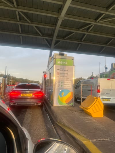 The image displays a multi-lane toll plaza on the M6toll, situated near Lichfield, United Kingdom. A dark grey BMW 740Li sedan, bearing UK registration TGZ 3474, is stationary in the leftmost visible lane, positioned directly in front of an automated toll booth. Its rear taillights are illuminated red. The toll booth, covered by an overhead grey metal canopy, features a sign with "M6toll" prominently displayed and instructions for drivers, including "stay in your vehicle," "no reversing," "CCTV in use," and options for contactless payment via card, phone, or smart device. A red signal light is active on the left side of the booth. To the immediate right, a white panel van with UK registration NA23 JWM is also stationary in an adjacent lane. A yellow plastic grit bin or utility box rests on the concrete kerb separating the lanes. The photograph is taken from inside another vehicle, with its front left section and wing mirror visible in the foreground. The sky above is light grey, suggesting either overcast conditions or early morning/late afternoon light. No individuals are visible outside of the vehicles.