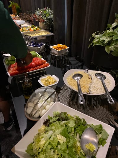 A salad bar setup featuring various options. A large square bowl holds green salad with a serving spoon. To its left, a bowl contains hard-boiled eggs and tongs. There are also dishes of sliced tomatoes, cucumbers, butter, and shredded cheese, accompanied by serving spoons. A bowl of tortilla chips is also present. The setup is on a black surface with greenery visible in the background.