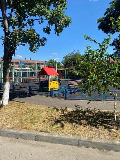 A brightly colored playground with a small red-roofed playhouse, a seesaw, and a swing set is visible under a clear blue sky on a sunny day. The playground is enclosed by a blue metal fence, and mature trees with green leaves surround the area. In the background, a building with a brick facade is partially visible behind a tall metal fence. The ground is covered with black rubber safety surfacing in the playground area, while the surrounding areas have dry, yellowed grass and asphalt pathways. A concrete curb runs along the edge of the asphalt in the foreground. The scene appears to be located in a residential or public park area.