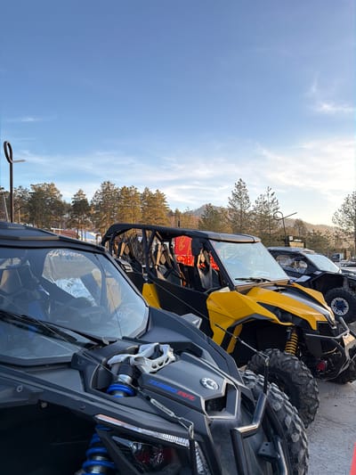 The image shows a row of three all-terrain vehicles (ATVs), presumably in the city of Озёрное, Russia. The ATVs are parked close together, with the front of each vehicle facing the viewer. The first ATV is dark gray, with "TURBO RR" branding visible. The second ATV is yellow, and the third appears to be black. In the background, there are trees, a building, a fence, and a utility pole. The sky is blue with some clouds.