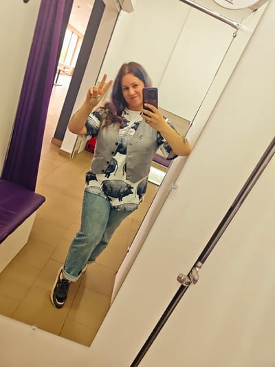 A woman is taking a selfie in a dressing room. She is wearing a gray vest over a shirt with a pig design, blue jeans, and sneakers. She is holding up two fingers in a peace sign. The dressing room has a purple curtain and a bench. A glimpse of a room with tables and chairs is visible through the curtain opening. The dressing room's interior is mostly white. There's no explicit evidence to confirm the location is in the city of Ильинское, Russia.