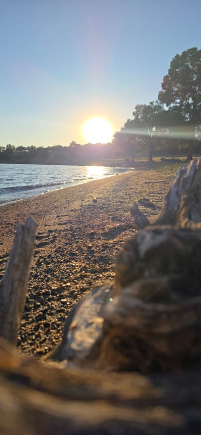 This is a vertical, eye-level, close-up shot of a pebbly beach and the edge of a body of water during sunset. The sun is low on the horizon, casting a warm glow across the scene. In the foreground, out of focus, is a piece of driftwood. The beach is composed of small pebbles, and the water's edge is visible with gentle waves. In the background, there is a treeline and a body of water, likely Issyk-Kul Lake in Kyrgyzstan. The sky is clear and blue, with the sun creating a bright glare and lens flare. The overall impression is one of a peaceful, natural landscape at the end of the day.