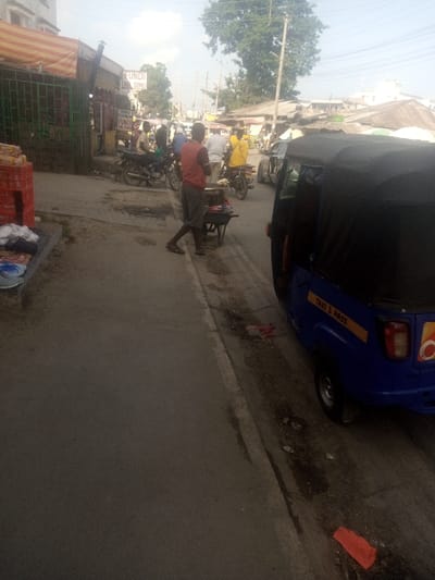The image displays a street scene within Mtopanga ward, Kenya, under a bright, hazy sky. In the immediate foreground on the right, a blue three-wheeled auto-rickshaw, marked "TAXI 3 PASS" in yellow lettering, is parked with its rear passenger door open. The ground adjacent to the vehicle and the curb consists of dirt and scattered debris, including red plastic remnants. A male individual, wearing a red sleeveless shirt and dark shorts, stands on the side of the road, facing left, positioned next to a dark-colored wheelbarrow containing assorted items. To his left, another individual is situated near a ground display of footwear, including sandals and slippers, arranged on a dark cloth.
In the midground, the road accommodates several motorcycles, some with drivers, indicating either stationary traffic or a slow-moving queue. One driver is notably clad in a yellow shirt, another wears a white helmet. On the left side of the frame, a commercial establishment is visible, featuring a red and white striped awning and green metal grating. Stacked red plastic crates are present near this establishment. Visible signs include "FUNDI WA SIMU" (Phone Technician). The background consists of various buildings, some with corrugated metal roofs, alongside a prominent large green tree and utility poles supporting overhead lines.