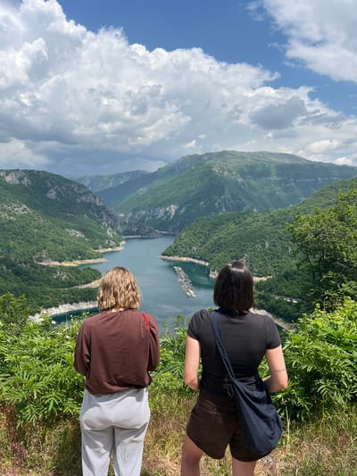 Two people are standing on a grassy hillside looking out over a lake or fjord surrounded by mountains. The person on the right is wearing a black shirt, brown shorts, and has a dark blue bag over their shoulder. The other person is wearing a brown sweatshirt and striped pants. In the background, there is a lake surrounded by mountains and a cloudy sky. There is a marina to the left of the lake. The location appears to be in the city of Seljani, Montenegro.