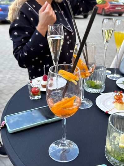 The image depicts a table set for an outdoor gathering, likely a celebration or a social event, with various drinks and small appetizers. In the foreground, a tall stemmed glass contains an orange-colored beverage garnished with orange slices and ice. To its left, another similar glass is partially visible. Behind these, a smaller shot glass holds a single red cherry in a clear liquid. A white plate with two small desserts, one topped with a raspberry and the other a small pastry, sits on the table. Further back, two tall flutes contain champagne or sparkling wine, and another glass is filled with a yellow beverage. A smartphone with a light green case is placed on the table. Two dark straws are prominent in the foreground glasses. The table is black, and the background shows a blurry outdoor setting with a red car and a person's upper body in a dark patterned dress. The scene is set in Zakharovo, Russia.