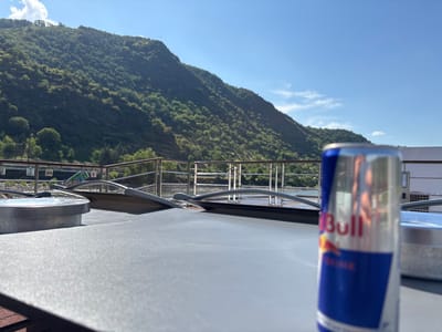 A close-up, slightly blurry shot captures a Red Bull can placed on a dark gray, textured surface in the foreground. The can is predominantly blue and silver with red lettering that partially reads "Bull." Behind the can, a metal railing runs horizontally, separating the foreground from a body of water and a lush green hillside. The hillside is densely covered with trees and vegetation, reaching towards a bright blue sky with a few wispy white clouds. The scene appears to be outdoors, possibly on a boat or a deck overlooking a river valley in Kestert, Germany, given the mountainous terrain and the presence of a river.