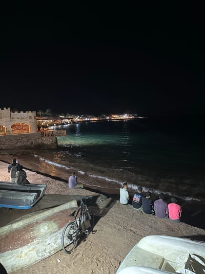 At night, a group of six people is positioned along the water's edge, bending down towards the sea. The people appear to be near a town. Nearby, a bicycle leans on an upturned boat. In the background, a pier lined with lights stretches out over the water. The location is Dahab, Egypt.