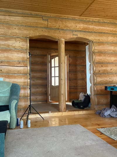 This appears to be an interior shot within a log cabin or house in the city of Пятница, Russia. The focus is on the log wall construction, showing a second-story opening or loft area. One small window can be observed near the middle of the frame. Random articles of clothing and other items appear to be resting near the edges. The orientation of the photograph is sideways.