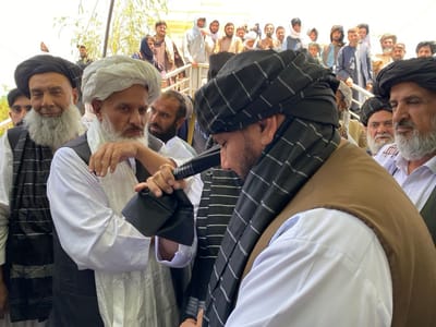 The image shows a group of men standing together in a crowded space. The men are all wearing traditional Afghan clothing, including turbans and long robes. One man in particular is standing at the center, wearing a black and white patterned turban and a brown vest, and is looking down. He is holding a black microphone in his left hand. The men are standing near a set of stairs. The scene is likely taking place in Zaker-E Sharif, Afghanistan, which is a prominent religious site.  The men appear to be engaged in a conversation or a religious ceremony.  A crowd of other men is visible in the background, standing on stairs and in the street.