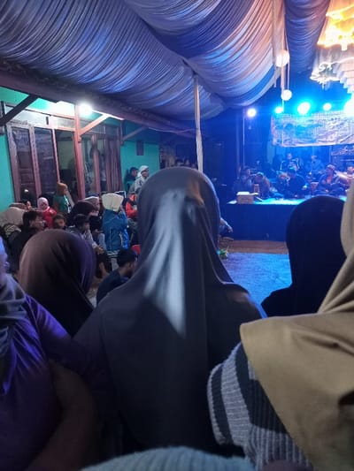 The image captures a nighttime community event or performance, viewed from behind the audience. A large number of people are gathered under a temporary white fabric canopy, which covers the area.
In the foreground, the backs of several individuals are visible, many of whom are women wearing hijabs, indicating they are part of the audience watching the event. To the left, a house with distinct green walls and brown wooden window frames serves as a backdrop, suggesting the event is taking place in a residential or village setting.
At the center and right, a brightly illuminated stage is the focal point, bathed in strong blue artificial light. On the stage, a group of musicians or performers can be seen with what appears to be musical equipment, suggesting a live band or traditional performance. Behind the performers, a banner is partially visible, featuring text that includes "LINGKUNG SINI PANG..." and "HARI JADI...", which translates to "anniversary" or "founding day," implying a celebratory occasion, possibly related to Pangalengan, Indonesia.
The overall ambiance is one of an engaged audience attending a public gathering or cultural event, characterized by the dramatic artificial lighting and the collective attention directed towards the stage.