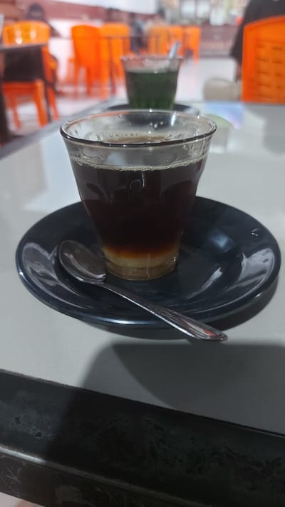 A clear glass mug with dark brown liquid and a lighter brown layer at the bottom sits on a black saucer. A silver spoon rests on the saucer, angled towards the mug. In the background, out of focus, are several bright orange chairs and tables, suggesting an indoor seating area. The scene is likely within a public establishment in Kruengmanyang, Indonesia.