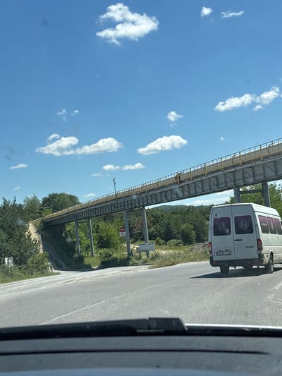 The image captures a view from inside a vehicle looking out at a highway and a raised conveyor belt system in Chavdar, Bulgaria. The sky is blue with scattered white clouds. A white van is driving on the right side of the highway. The van has a license plate reading "CB 9817 OB" and several circular logos on its rear windows. The conveyor belt system, supported by metal poles, stretches across the scene, elevated above the roadway and surrounded by trees and greenery. A dirt road slopes upwards to the left of the highway, disappearing behind trees. A traffic sign with a red circle and a horizontal bar is visible on the right side of the conveyor belt supports. The overall impression is a rural or semi-industrial landscape under a bright sky.