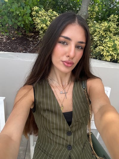 In an outdoor setting, a young woman with long, dark brown hair and brown eyes is taking a selfie. She is wearing a dark vest with white pinstripes over a black top. She has several necklaces on, including one with a cross pendant and another with a rectangular pendant. Her left arm is visible, and she is holding a dark green purse with a gold chain strap over her right shoulder. Behind her, there are lush green and yellow-green plants, and part of a tree trunk is visible on the right. The background also shows a white ledge and white chairs, suggesting a patio or outdoor seating area in Hallandale Beach, United States of America.