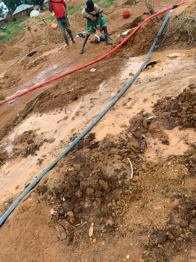 In Buga da Gindi, Nigeria, a rural landscape displays evidence of mining activities. Two individuals are visible in the background, one wearing a red shirt and jeans, the other in a green and white shirt with green shorts. The ground is a mix of mud and dry soil, with piles of excavated earth. Two large hoses, one red and one blue, run across the scene, suggesting water is being used in the mining process. The blue hose lies partly submerged in a muddy puddle, with a dark object, possibly clothing, also in the water. Scattered debris, including a plastic bottle, is present on the muddy ground. The overall scene is one of active, albeit rudimentary, resource extraction in a natural, unpaved environment.