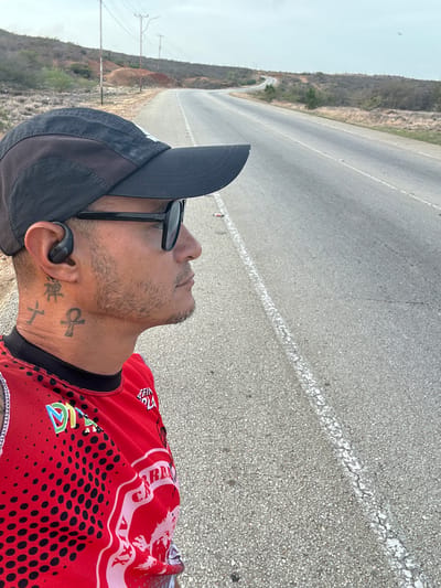 The image shows a man wearing a black baseball cap, sunglasses, and a red shirt with black polka dots and white text. He has a tattoo on his neck and is wearing an earbud. The man is standing on the side of a paved road that curves into the distance. The landscape is dry and hilly, with sparse vegetation and utility poles visible. The sky is overcast. The road appears to be in a rural or semi-rural area, possibly on the outskirts of La Guaira, Venezuela.