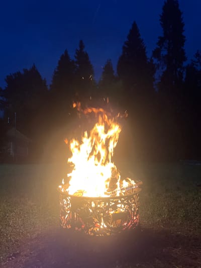 A fire is burning in a decorative metal fire pit in a grassy yard at dusk. The fire pit has intricate cutouts that allow the flames to be visible. In the background, silhouetted pine trees stand against a dark blue sky. To the left of the fire pit, a building with a pitched roof and a porch light is visible. The scene is illuminated by the bright flames of the fire, casting a warm glow on the surrounding grass. The image does not provide any specific details or indicators that would allow for the identification of its location within the city of Popovka, Russia.