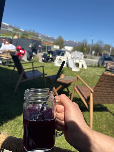 A hand holds a glass jar filled with a dark red liquid and a black straw. The jar is on a table, and there is a person sitting in the background with motorcycles parked nearby. The scene appears to be outdoors in Pirdop, Bulgaria, given the visible mountainous landscape and the presence of vehicles.