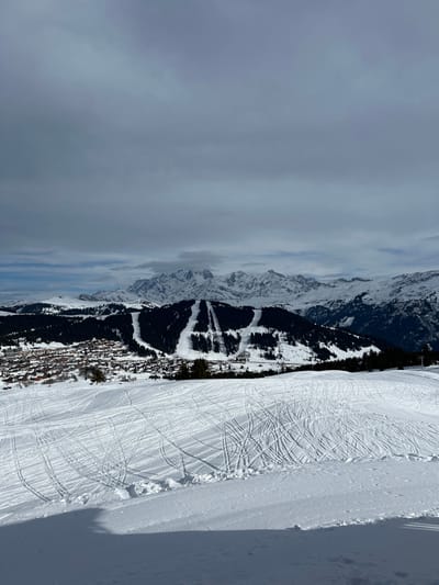 The image displays a wide, panoramic view of a snow-covered mountainous landscape in Villard-sur-Doron, France, under an overcast sky. In the foreground, a gentle snow-covered slope is visible, extensively marked with numerous intertwined ski tracks. The midground features a prominent, densely forested mountain with several wide, treeless ski runs descending its slopes. To the left, at the base of this forested mountain, the village of Villard-sur-Doron is discernible, composed of numerous buildings with dark roofs, partially obscured by snow and trees. In the background, a vast range of rugged, snow-capped mountains stretches across the horizon under a predominantly grey, cloudy sky. No individuals or active subjects are present in the scene.