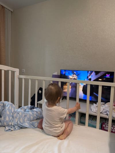A young child is sitting in a crib, facing away from the camera, and watching television. The child is wearing a white t-shirt and appears to be a toddler. The crib is white with wooden bars. A blue and white cloud-patterned blanket is in the crib with the child. A television is mounted on a shelf to the right of the crib, displaying a colorful image. The room appears to be a bedroom. The location is the city of Шихово, Russia.