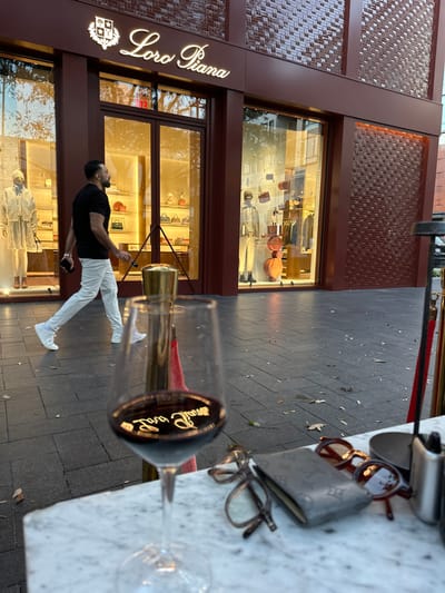 A man in a black t-shirt and white pants stands in front of a Loro Piana store, holding a phone. The store's windows display various bags and clothing. A wine glass with the Loro Piana logo and a small amount of red wine sits on a marble table in the foreground. Several pairs of eyeglasses and a Louis Vuitton wallet also sit on the table. The photo is taken from an angle, with the sidewalk pavement visible behind the table.