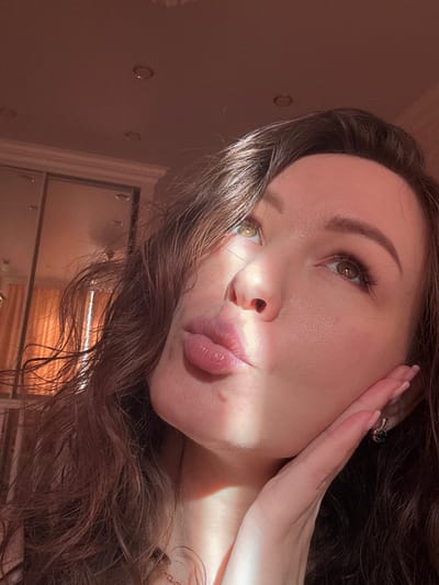 A close-up, angled portrait features a young woman with long, wavy dark brown hair. She is captured in soft, direct sunlight, creating strong highlights and shadows across her face. Her head is tilted, and her gaze is directed upwards and slightly to the left. Her lips are slightly puckered in a pouty or kiss-like expression. Her right hand is gently touching her cheek, displaying a ring with a dark, possibly green, stone on her ring finger, and her nails are painted a light nude color. A small earring with a dark gemstone is visible on her right earlobe. Her eyes are a warm brown or hazel, accentuated by long eyelashes and sculpted eyebrows.

The background is softly out of focus, revealing an indoor setting with a warm, light brown or beige wall. Subtle circular elements are visible on the wall, possibly indicating decorative studs or recessed lighting. On the right side of the frame, reflections in a mirrored or glass surface show hints of the room's interior, including what appears to be warm-toned surfaces, possibly wood. The strong, low-angle light suggests it is either morning or late afternoon. No text is visible in the image. The setting is an indoor location in Yuzhny, Russia.