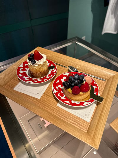 An overhead, slightly angled shot captures two elegantly presented desserts on a wooden serving tray, set within what appears to be a luxury retail environment or high-end café. The scene is likely indoors, possibly within an Hermès boutique, supported by visible branding.

The top dessert is a vibrant fruit tart, generously topped with an assortment of fresh berries including bright red strawberries, deep blue blueberries, red raspberries, and dark blackberries. A small pink macaron and a gold-colored serving utensil are also part of its presentation. This tart rests on a distinctive round plate featuring a red and white swirling, organic pattern, with "HERMÈS PARIS" subtly inscribed on its rim. Below the plate is a small, off-white linen napkin embossed with an "H" logo.

The second dessert, positioned slightly below and to the left, is a smaller pastry, possibly a scone or mini cake, adorned with a swirl of white whipped cream and garnished with a single strawberry, a few blueberries, and raspberries. This dessert is also served on an identical red and white patterned plate with "HERMÈS PARIS" text, and a matching "H"-emblazoned napkin. A silver spoon is visible near the upper dessert, and another silver utensil is partially seen near the lower one.

The wooden tray itself is light-colored, with a fine grain. Surrounding the tray are reflective surfaces and glass, suggesting a display case or a polished table. In the background, dark teal or green surfaces are visible, along with glimpses of other displays, including a clear glass case through which a large, stylized pink "H" can be discerned. The overall presentation suggests a refined break or snack experience in Zapopan, Mexico.