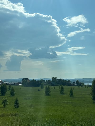 A wide-angle, eye-level shot captures a rural landscape under a dynamic sky, likely taken in Ir'yak, Russia. The foreground is dominated by a vast, undulating expanse of lush green grass, dotted with scattered trees of varying sizes and densities. In the middle ground, a cluster of buildings, suggesting a village or farmstead, is nestled amongst a dense line of trees. The architecture appears rustic, with at least two prominent structures featuring dark roofs and what could be wooden siding. The background reveals a hazy horizon line, with subtle hills and a body of water, possibly a lake or river, visible in the distance. The sky is a dominant feature, filled with a mix of bright blue sky and large, billowing white and gray clouds. Shafts of sunlight pierce through the clouds, creating dramatic lighting effects across the scene. There are no people or animals visible in the frame.
