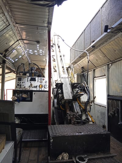 The image depicts the interior of a mobile drilling rig, likely situated in Raygorodok, Kazakhstan. The scene is industrial and functional, with a focus on the machinery. A large drilling apparatus dominates the center, with its mast extending upwards. Various hydraulic hoses, pipes, and mechanical components are visible, painted in a mix of white, grey, and yellow.
To the left, a control panel is mounted, featuring multiple gauges, dials, and switches. This panel is white and appears to be the operational hub of the drilling rig. Below it, a white housing contains what seem to be circuit breakers or control switches.
The rig is housed within a structure that resembles a large, elongated shed or trailer, with corrugated metal walls and a sloped, reflective metal ceiling. The interior lighting is provided by strips of LED lights running along the ceiling. In the background, a window with a bright, washed-out view suggests it is daytime.
On the right wall, a rack of wrenches and tools is neatly organized, indicating a workspace where maintenance and operations are performed. The floor appears to be made of wooden planks, showing signs of wear and tear. In the foreground, a textured metal platform sits in front of the drilling equipment, likely a standing area for operators. A metal tool, possibly a wrench or clamp, lies on the floor in the lower right corner. There are no people visible in the image. The overall impression is one of a working industrial environment.