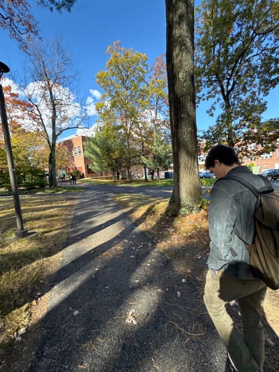 This image depicts an outdoor scene on a sunny autumn day in Wellesley, United States, likely within a college campus or institutional grounds. In the foreground, a male individual is seen from the mid-back and right side, walking on a dark paved pathway. He has dark, curly hair and is wearing a dark gray long-sleeved top and olive green pants, with a brown backpack slung over his right shoulder. His head is slightly bowed. The pathway shows numerous long, distinct shadows from trees, indicating strong sunlight, and is scattered with fallen autumn leaves. To the left, a dark utility pole stands upright.

The midground features grassy areas, more of the paved pathway, and several mature trees displaying various stages of autumn foliage, with leaves ranging from green to yellow, orange, and red. In the background, a two-story brick building with multiple windows is prominent. Near this building, a small group of approximately three to four indistinct figures appears to be walking on another path. Further to the right in the background, additional trees with autumn colors are visible, and behind them, parked cars indicate a nearby road or parking area. The sky is clear blue with sparse white clouds.