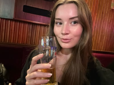 A young adult female is depicted indoors, seated, holding a glass of beer. She has long, light brown hair, fair skin, and light-colored eyes, looking directly at the camera with a subtle smile. She wears a dark-colored top, and a delicate necklace is visible. Her right hand holds a pilsner-style glass containing a light-colored beverage, likely beer, with the word "PREMIUM" partially visible on the glass. The background consists of a reddish-brown wooden slatted wall, a dark red padded bench or booth below it, and a dark horizontal fixture above the wall. The lighting is warm and dim. This scene is located in 마포구, South Korea.