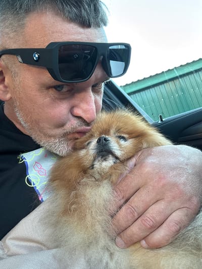 A man with graying hair and stubble, wearing black sunglasses with a BMW logo, is holding a small, fluffy tan dog. The man is leaning in to kiss the dog's head, and the dog appears relaxed. They are inside a vehicle, with the car door visible in the background. The environment outside the car is out of focus, but appears to be an outdoor setting with a green roof structure. There is a patterned cloth draped over the man's arm and under the dog. The image is a selfie taken by the man. The location is not specified.