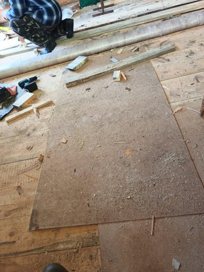 In Islambaevo, Russia, a person wearing a blue and black plaid shirt, camouflage pants, and black boots kneels on a wooden floor covered with sawdust and scattered wood scraps. The person is near a workbench area where a cordless drill, tools, and pieces of wood are visible. A large piece of pressed wood or particle board lies on the floor in front of the person, also covered in sawdust. The environment appears to be an indoor workshop or construction site, with unfinished wooden planks forming the floor and walls. The scene suggests ongoing construction or renovation work.