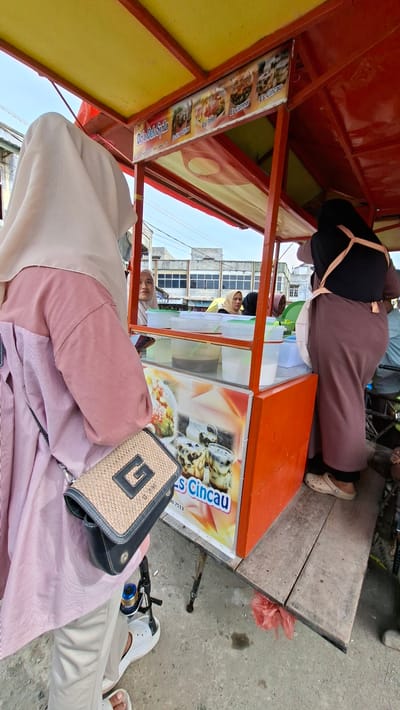 In this media file, the scene captures an outdoor setting with a focus on a food cart or stall. Two individuals are partially visible, one wearing a pink headscarf and pink striped shirt, and another wearing a black top and a muted pink skirt or dress. The food cart is brightly colored in red and yellow, featuring a display of drinks and food items. Visible on the cart are signs with text in Indonesian, including "NAVA KIDS," "Sirup Cincau," "Es Teler," "Es Campur," and "Es Cincau." A woven handbag with a prominent "G" logo sits on the lap of the person in the foreground. In the background, buildings with windows and a glimpse of the sky suggest an urban or semi-urban environment. The ground appears to be paved or concrete. The lighting suggests it is daytime, and the overall atmosphere seems to be casual and possibly commercial, with the food cart indicating a point of sale for refreshments.
