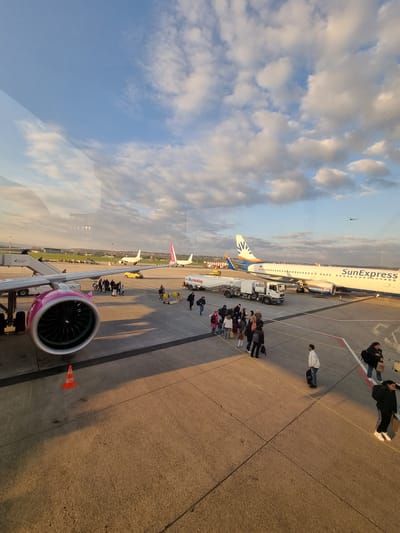 The image is taken from inside an aircraft or airport terminal, looking out onto an airport apron in Echterdingen, Germany. The sky is partially covered with clouds. A SunExpress aircraft is parked on the apron, and passengers are boarding or disembarking. There are several other aircraft parked in the background, including one with a pink tail. A jet bridge with a pink engine is visible in the foreground, presumably connected to the aircraft where the photo was taken. Airport ground support vehicles are visible near the SunExpress plane. Groups of people are standing around, likely waiting to board or having disembarked from a plane. An orange cone is near the engine.