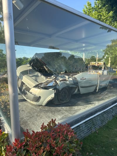 The image displays a severely damaged light-colored car, identified as a Renault Megane, exhibited within a large glass enclosure outdoors. The car's front end is heavily crushed and deformed, with parts of the engine compartment exposed, and the windshield is shattered. The body panels show significant impact damage, and the wheels appear to be damaged or missing tires.

The glass display case reflects the surrounding environment, which includes lush green trees and a clear blue sky, suggesting a sunny day. Reflections of a distant food cart or kiosk, with text like "SHAWARMA" visible, are also seen in the glass. Adjacent to the display, there is a vibrant red-leafed bush and a metal grate on the ground. The ground around the enclosure is paved or concrete.

The car is presented as a static display, likely serving as a stark visual reminder of the consequences of traffic accidents, often used in public road safety campaigns. The visible text on the car explicitly spells out "RENAULT" on the front bumper and "MEGANE" on the side panel. The bright sunlight and distinct shadows indicate it is daytime. The scene is located in Esenler, Turkey.