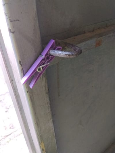 The image shows a purple clothespin attached to a rusty metal ring. The clothespin is made of plastic and has a metal spring. The metal ring is circular and appears to be attached to a larger metal structure. The background is a gray, textured surface. The scene is outdoors, possibly in a city like Gori, Georgia, given the industrial-looking metal structures. There are no people or animals visible in the image. The lighting suggests it is daytime.